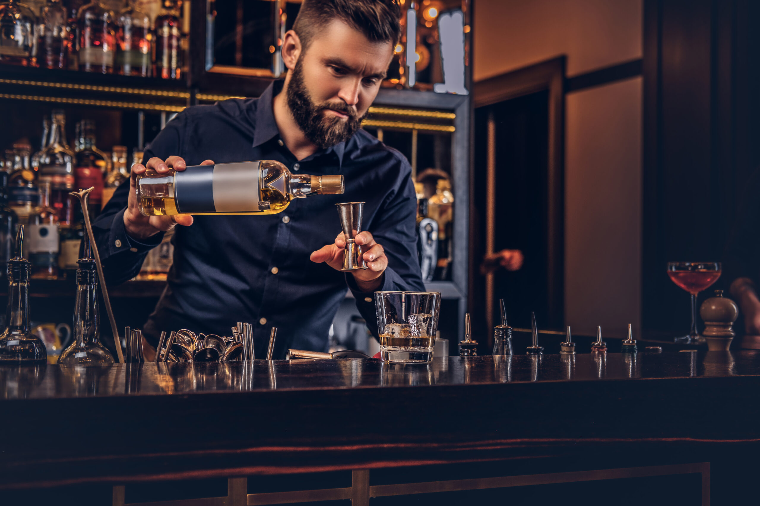 Stylish brutal bartender in a black shirt makes a cocktail at bar counter background.